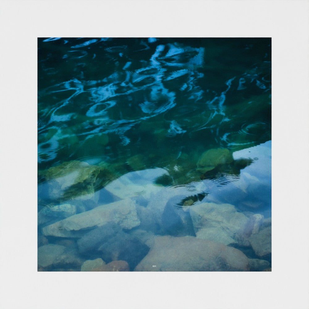 A square photographic print depicting a close-up view of clear blue-green water over smooth rocks, surrounded by a wide white border.