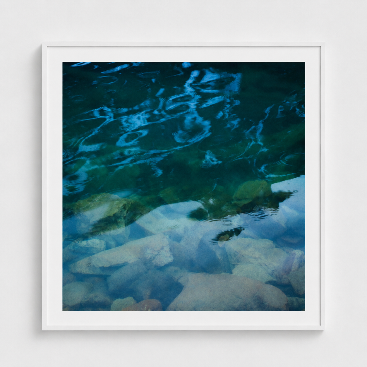 A framed square art print depicting clear blue and green water over smooth stones, presented in a white frame against a light gray wall.